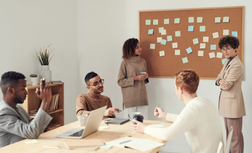 3 professionals sitting at a conference table looking at 2 other professionals who are standing by a board covered in stickie notes.