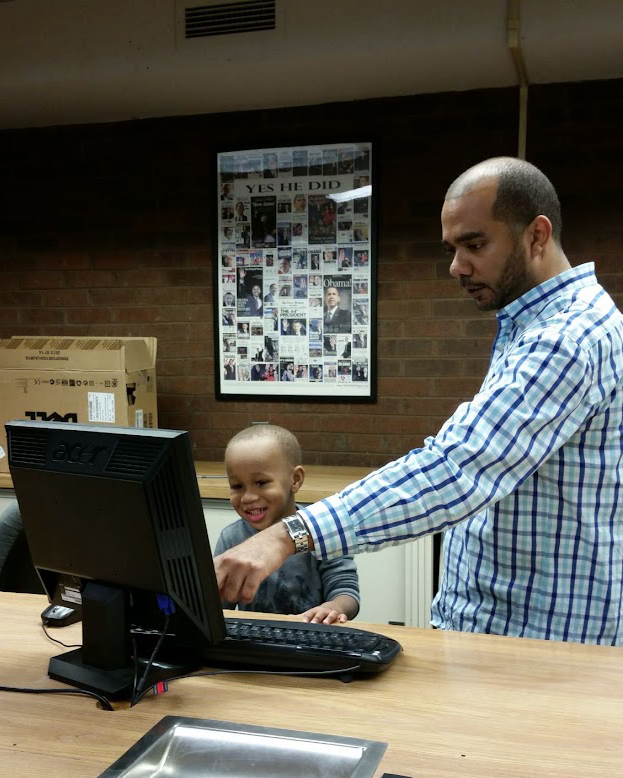 A man points to a computer screen while a child sits and watches.